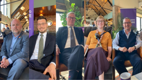 A row of people, four men and one woman, sitting on a sofa with microphones in front of them. They are looking at the camera and dressed smartly.