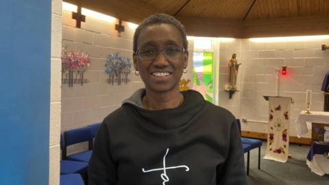 A woman smiling into the camera she has short brown hair and is wearing glasses. She has a black jumper on with a white cross in the background. There is the inside of a chapel behind her and we can see blue chairs and a podium which has a cross on it too.