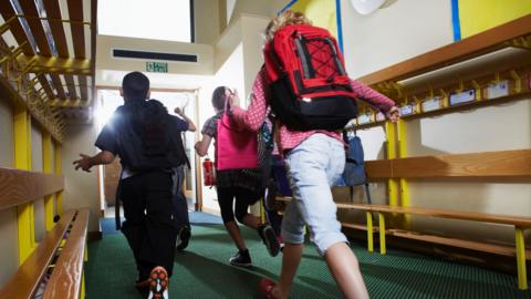 Children running down a corridor inside a school towards an open door where sunlight pours in. On the walls either side of the corridor are wooden benches and coat hooks.