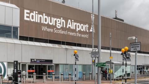 A view of the entrance to Edinburgh Airport. Underneath the name of the airport is a slogan reading Where Scotland Meets The World. As well as the entrance, there are shelters visible, along with a sign saying Assembly Point 1.  