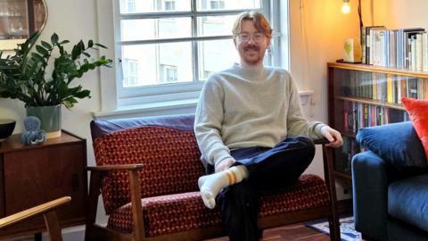 Matt McCallum sits on a bright orange and red subway seat converted into a couch with wooden armrests and legs. He is in a living room by the window. He has ginger hair and is wearing a grey jumper, navy trousers and white socks.