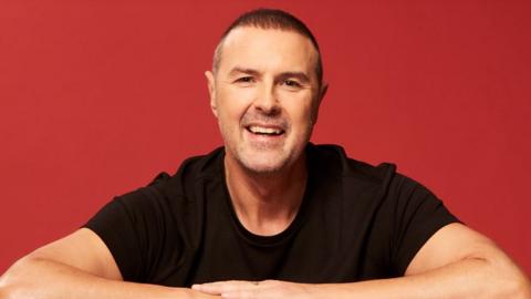 Paddy McGuinness smiling at the camera while leaning on a table with his arms folded in front of him, against an orange/red background. He has short dark hair and is wearing a black t-shirt.