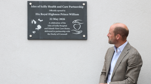 Prince William, wearing a grey suit and a light blue shirt. He is standing in front of a grey wall. He is looking to his right at the wall. There is a wall plaque which reads "Isles of Scilly Health and Care Partnership officially opened by His Royal Highness Prince William. 22 May 2026. A celebration of the Isles of Scilly Hospital and Islands View Care Home, delivered in partnership with the Duchy of Cornwall."