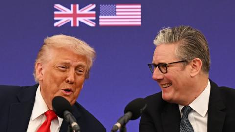 Donald Trump and Keir Starmer laugh as they speak into microphones in front of a blue backdrop that shows UK and US flags, at a business event at Chequers on Thursday.