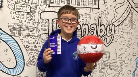 Riley is a boy with short hair, glasses and a blue hoody. He is holding a pen and a book of raffle tickets in his right hand. In his left is a red and white football containing the logo for Castore, the Middlesbrough badge and lots of signatures. Behind him is a huge drawing of a map of Thornaby.