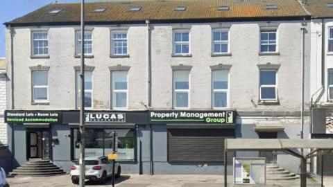 A Google Streetview image looking towards a dark white and grey building. It is three storeys in height and has black doors and shutters on the first floor belonging to a property business.