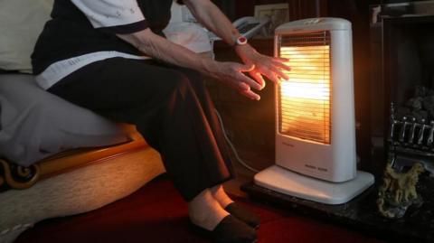 A white portable electric fire with a glowing bar inside, placed in front of a fireplace.  The hands of an elderly woman, who is sitting on a sofa, are seen stretching out towards the fire.