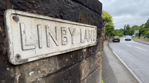 Street sign of Linby Lane, in Linby, Nottinghamshire, with two cars on the road to the right.