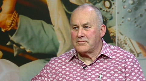 Dave Land is sitting down and wearing a pink and white patterned shirt. He has a microphone attached to his shirt and he is looking to the left of the camera. 