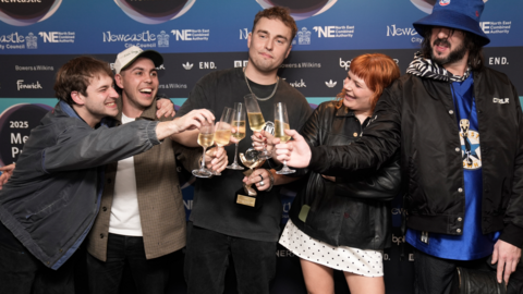 Sam Fender and his band clinking champagne flutes after the award win. Fender is holding his trophy as well as his own glass.