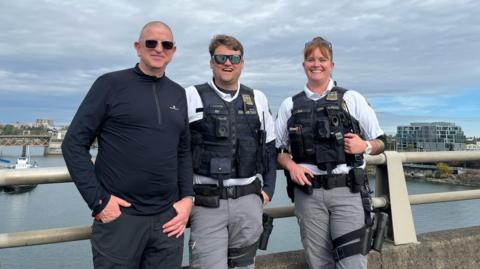 A man wearing sunglasses and a dark-coloured top is standing next to two police officers wearing flak jackets. They are leaning against railings above a body of water with boats and buildings 