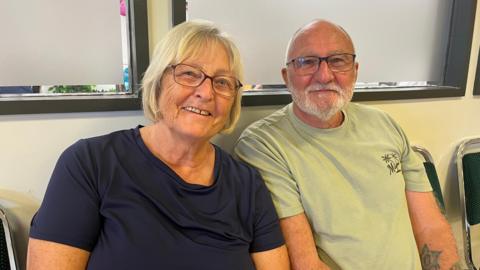 A head and shoulder shot of a woman and a man - Bea and Frank Carter - sitting down smiling at the camera. Bea has blonde, bobbed hair and is wearing glasses and a navy t-shirt. Frank is bald and has grey facial hair and is wearing glasses and a green t-shirt. He has a tattoo of a star on his left arm.