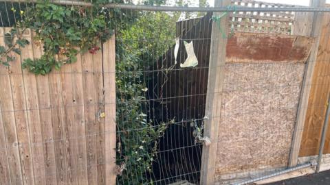 The entrance to a footpath, with overgrown brushes and a gate closing it off. The remnants of a closure sign can be seen tied to the gate. 