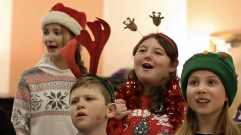 Members of the children's choir - three girls and a boy - singing, in Christmas jumpers and hats