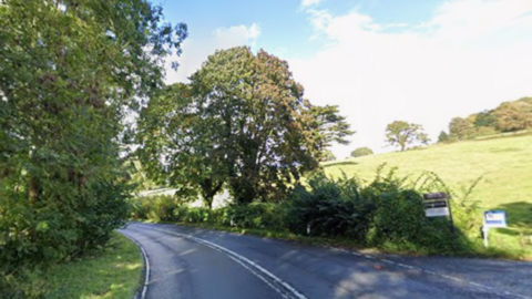 A country A-road sweeps around a corner, bordered on each side by hedges and some trees. To the right of the road is a lane with some "For Sale" signs on them, and there is a field sloping down to the road from the right. It is a sunny day.