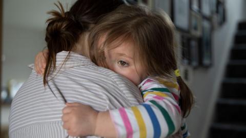 A woman with brown hair tied up and wearing a grey and white striped shirt hugs a young girl who is wearing a colourfully striped long-sleeved top and has her brown hair tied in pig tails