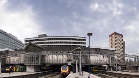 An exterior view of Birmingham New Street station showing multiple tracks and platforms side by side. There are three trains in view on the tracks, with a high rise building to the right in the background