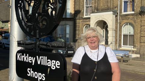 Wendy Dalton, wearing glasses, stands beside a black sign reading "Kirkley Village Shops" with black plastic wrapped around an arrow pointing to shopping area