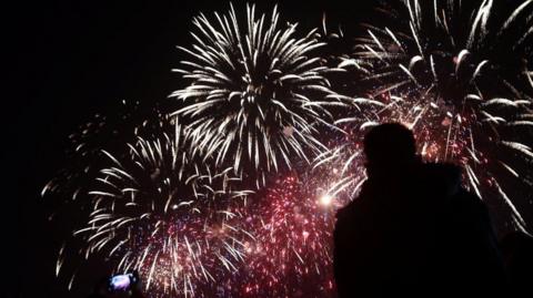 Red and white star-shaped fireworks in a black sky. The silhouette of a person is in the foreground with their face turned up watching the display and there is a hand holding a mobile film.
