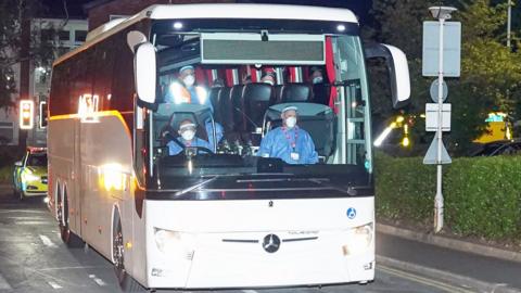 The front of a coach carrying 20 Britons to Arrowe Park Hospital. Sitting at the front are three people wearing personal protective equipment including face shields, face masks, gloves, and coveralls. The tops of some passengers heads wearing face masks can be seen.