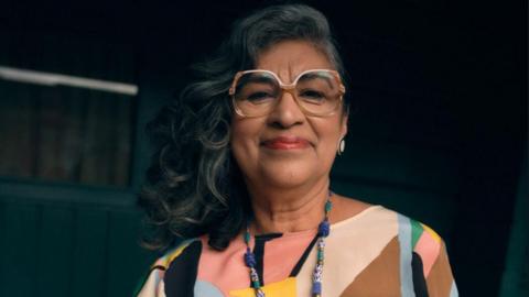 A woman with long dark hair, large glasses and a colourful patterned top in front of a dark background