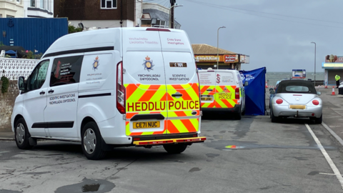 The end of Mackworth Road, leading to the beach and the sea at the end. 
There is a silver car parked with police vans beside it. A restaurant can be seen in the background. 