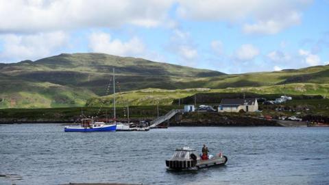 A view of the isle of Ulva, showing a ferry near the dock. A couple of buildings, including a pub/restaurant, can be seen, with rolling hills behind them.
