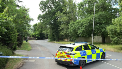 A police car and police tape marking off a road following a crash. A police officer can be seen far in the distance conducting investigations.