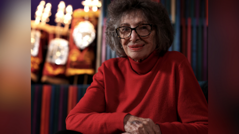 A landscape image of Holocaust survivor Dorit Oliver-Wolff at a synagogue in Brighton. Dorit has her right hand over her left and is wearing a red jumper and brown-framed glasses.