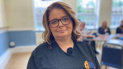 A head-and-shoulders portrait of Karla Rutherford. She is at the community centre where the support group meets and the blurry background shows people sitting down in a circle. Ms Rutherfod is wearing a black t-shirt with a logo. She has shoulder-length, dark blond wavy hair and wears glasses.