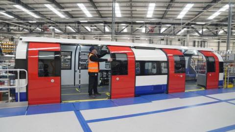 A blue, red and white train carriage inside a factory. The two sets of red doors of the carriage are open. A man wearing an orange hi-vis vest is stood in the doorway of the left set of doors, with his hands holding on to the side of one of the doors.