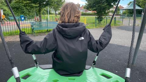 An anonymous boy with his back to the camera in a park. He has brown hair down to his neck and is facing away from the picture. He's on a swing, wearing headphones, and is aged about 12. This is a stock picture. 