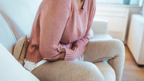 A woman sitting on a cream sofa, with her arms folded around her stomach. the woman's face cannot be seen. She is wearing a pink jumper and beige leggings and is partly hunched over.