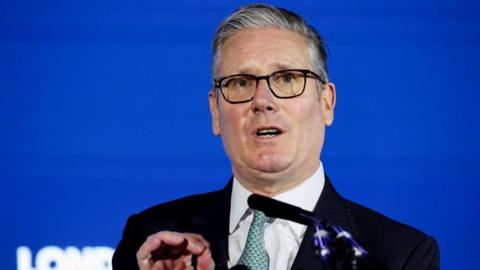 Sir Keir Starmer is stood in front of a blue background and a microphone is visible in front of him. He is wearing a dark coloured suit jacket, white shirt, a patterned tie and glasses.