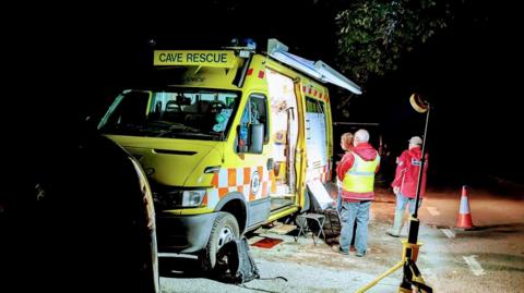 Derbyshire Cave Rescue Organisation van with volunteers outside during the evening. It is a van with decals similar to an emergency service vehicle in yellow with red and white checked bands. 