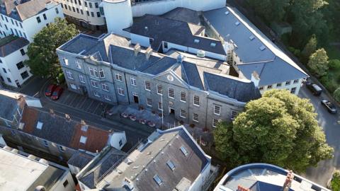 An aerial view of the States of Guernsey building. It is a large grey stone building, surrounded by streets and trees.