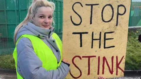 A young woman with long blonde hair, in a grey coat and yellow high vis vest, holds a sign that says 'stop the stink' in front of large green skips at a landfill site.