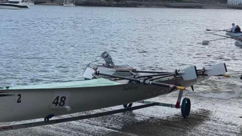 A swift style rowing boat on the slip way above the water. It is long with a number of ores inside it. There are some rowers in another boat in the background along with some larger boats on the water. 