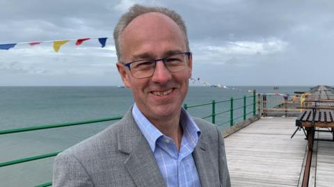 Tim Baker wearing glasses, a blue shirt and grey suit jacket, smiling while standing on the Queen's Pier in Ramsey on a cloudy day.