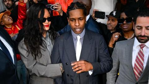 A$AP Rocky locks arms with his partner Rihanna outside an LA courthouse as crowds of people and reporters surround them with cameras. 