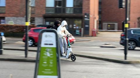 Two people ride together on an e-scooter along a city street. The rider in front wears a light-colored hoodie and grey joggers, while the person behind wears a dark jacket with the hood up. 
