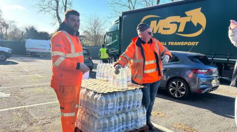South East Water crew members at Tunbridge Wells Sports Centre on St John's Road, Tunbridge Wells. A temporary water station has been set up there. Both crew members are wearing orange high vis and are stood beside two crates of water bottles. 