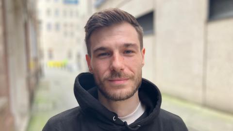 A close up of Tyler with an alleyway and buildings in the background. He has a brown beard and short brown hair and he is wearing a black  hoodie with a white t-shirt underneath. 