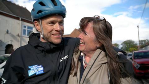 A man with a blue cycle helmet and black top hugging a woman in a grey coat with long brown hair
