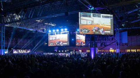 A huge crowd of spectators fills a darkened exhibition hall, silhouetted against bright blue and white stage lights. Giant screens above the stage display a first-person shooter game in progress, with scoreboards visible at the top of the display. The industrial ceiling of the NEC is visible above, with rigging and spotlights cutting through the haze.