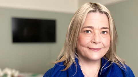 Harriet Tyce, a woman with blonde hair standing inside a room with grey walls. She is wearing a blue outfit and is looking directly at the camera and smiling.