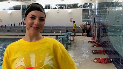 A woman in a yellow t-shirt and a black swimming hat standing and smiling in front of an indoor swimming pool.