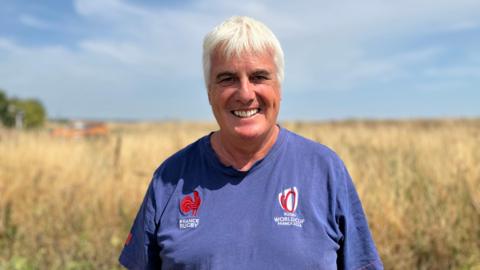 Tim Carson smiles at the camera on a sunny day, a blurred field and blue sky behind. He is wearing an old blue rugby world cup T-shirt.