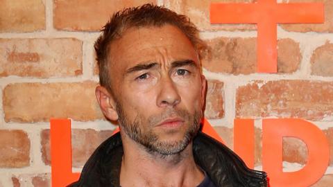 Close-up of James Holder, a small man with grey facial hair, dark grey hair and tanned skin. He is in his 50s and is standing in front of a brick wall with orange writing on it as he poses wearing a collared leather jacket.