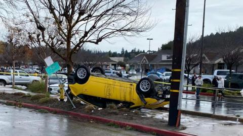 A yellow five-door car that has been flipped upside down and onto the pavement by a tornado in Scotts Valley. A shop and a large car park with other parked cars can be seen in the background.